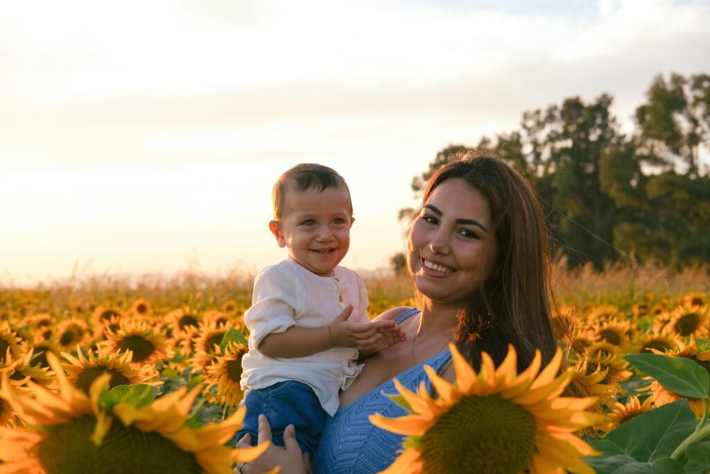 A smiling mother holds her baby surrounded by vibrant sunflowers at sunset.